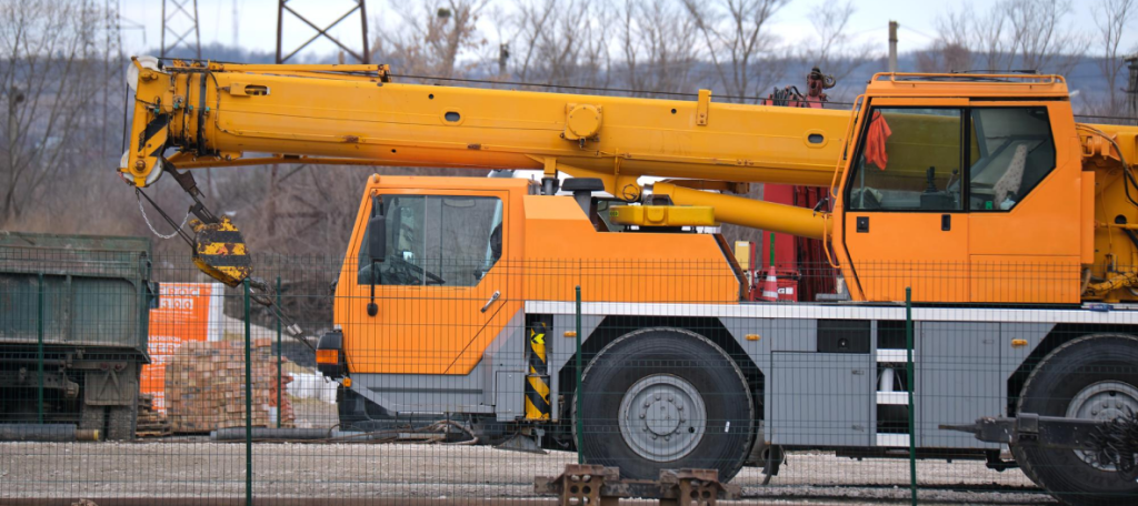 Side profile of a yellow all-terrain mobile crane parked on an industrial construction site for taxi crane services.